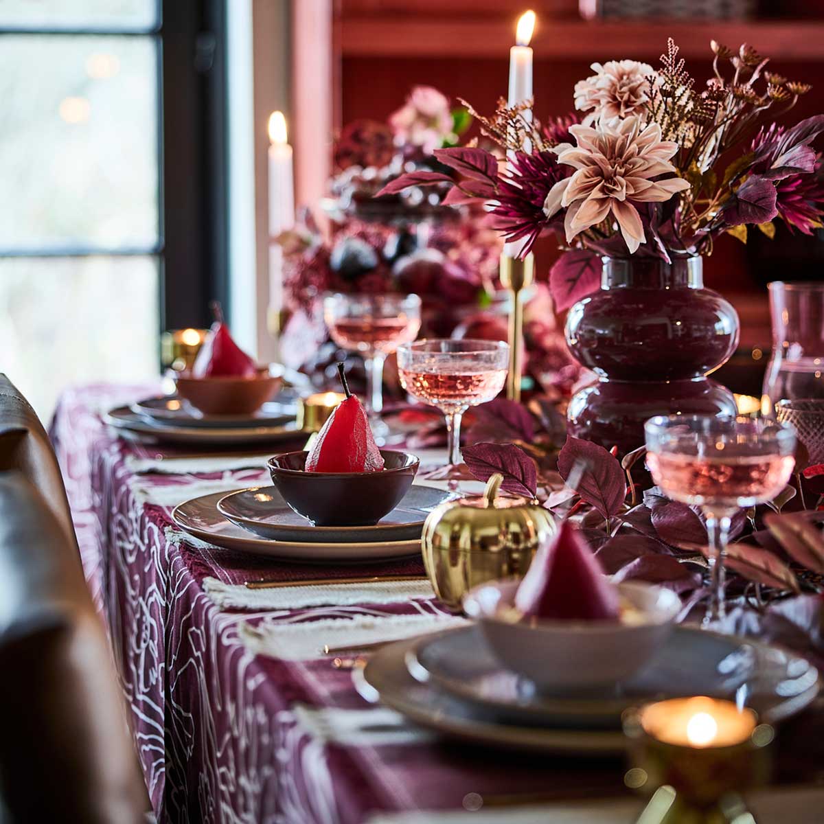 A pink and red table setting with lit candles, HÖSTAGILLE artificial bouquets in vases and poached pears in bowls.