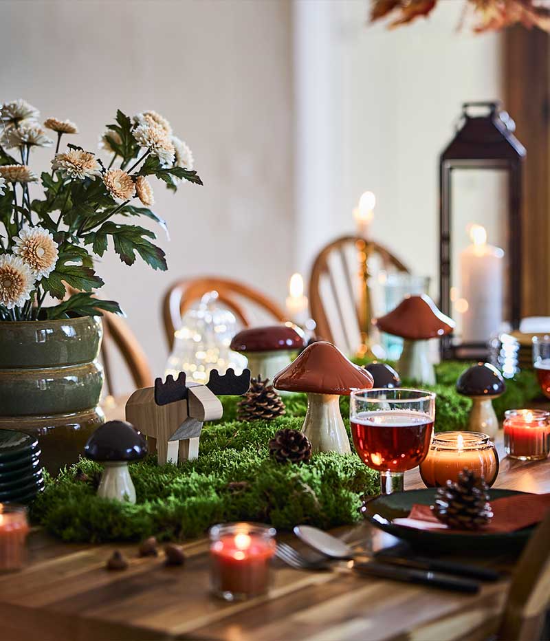 A woodland-themed dining table is set with HÖSTAGILLE decorative mushrooms and moose, lit candles, moss and pinecones.