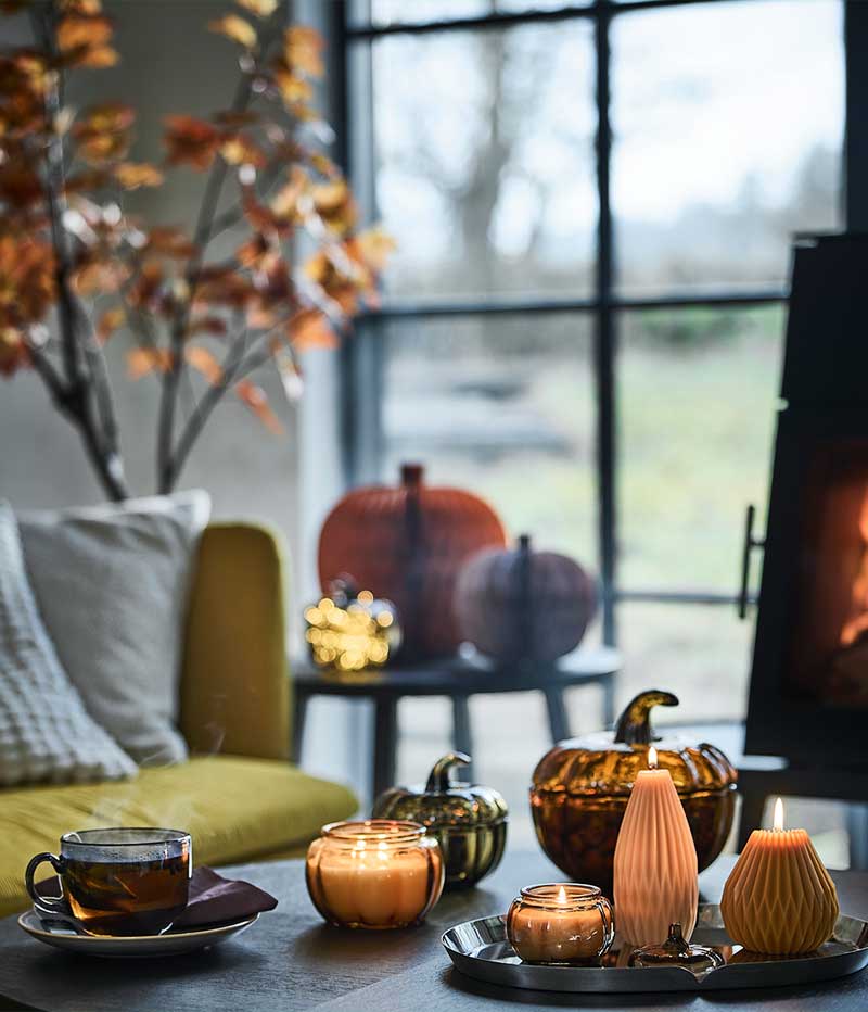 A cosy corner next to a window has a coffee table with lit HÖSTAGILLE scented candles, pumpkin-shaped pillar candles and mug.