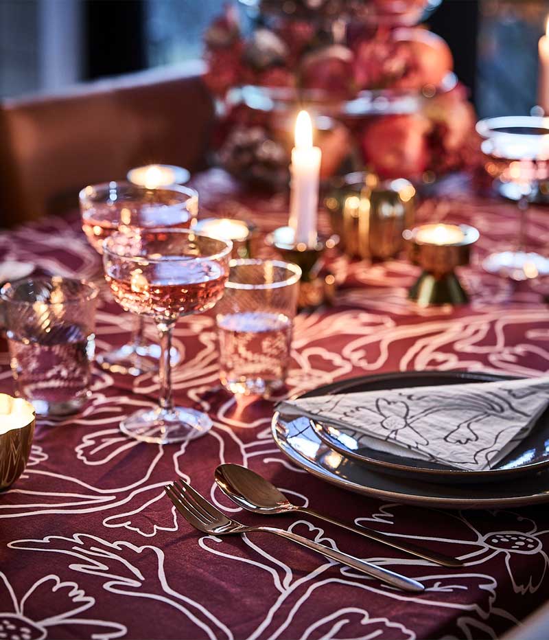 A pink and red table dining table is set with lit candles, a red patterned HÖSTAGILLE tablecloth, paper napkins and coupes.
