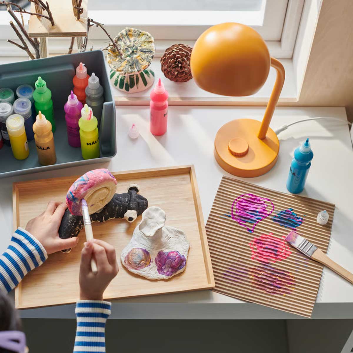 A bright room with a small desk with a yellow KORVSNÖRE lamp and paint bottles. A child paints a snail on an OSTBIT tray.