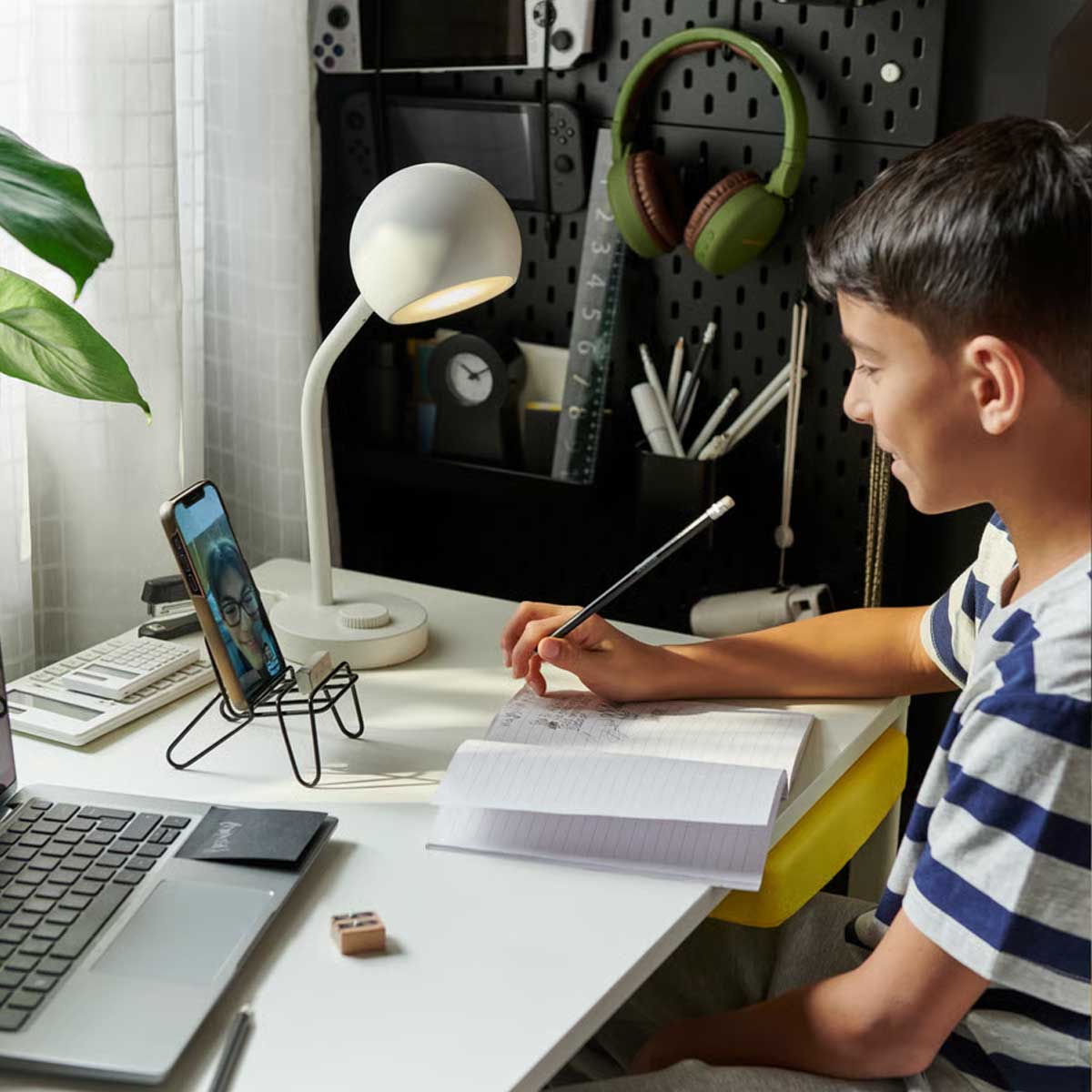 A young boy sits at a white ENKROKEN desk, with a laptop, notebook, and a white KORVSNÖRE LED lamp on top.
