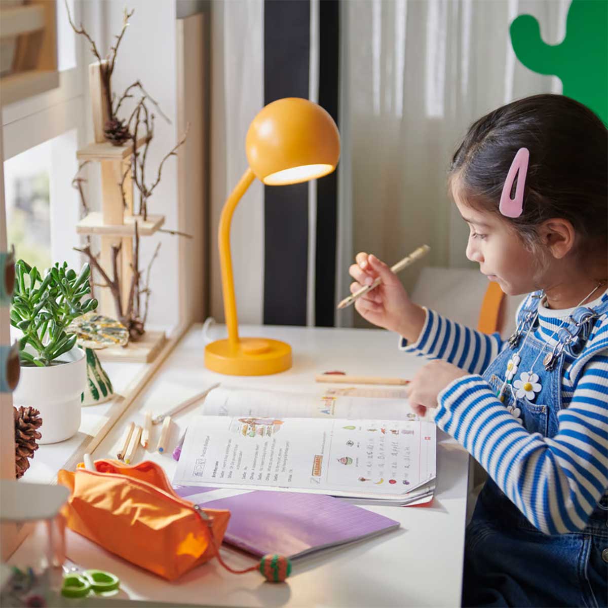 A child with pen in hand sits at a white desktop doing homework. An orange KORVSNÖRE LED lamp provides illumination.