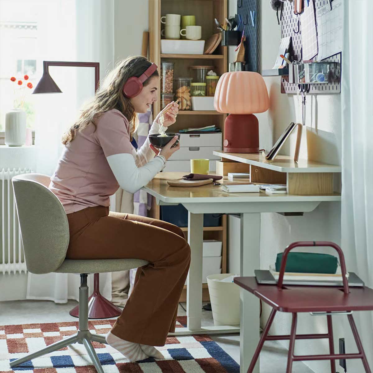A young woman sits in a dark beige KRYLBO swivel chair at a desk, eating noodles, surrounded by storage and decorative items.