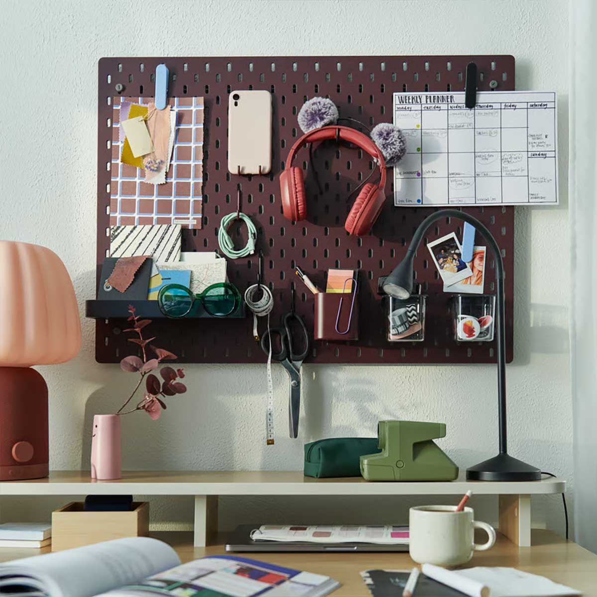 A study space features a dark red SKÅDIS pegboard above a desk, displaying stationery and headphones.