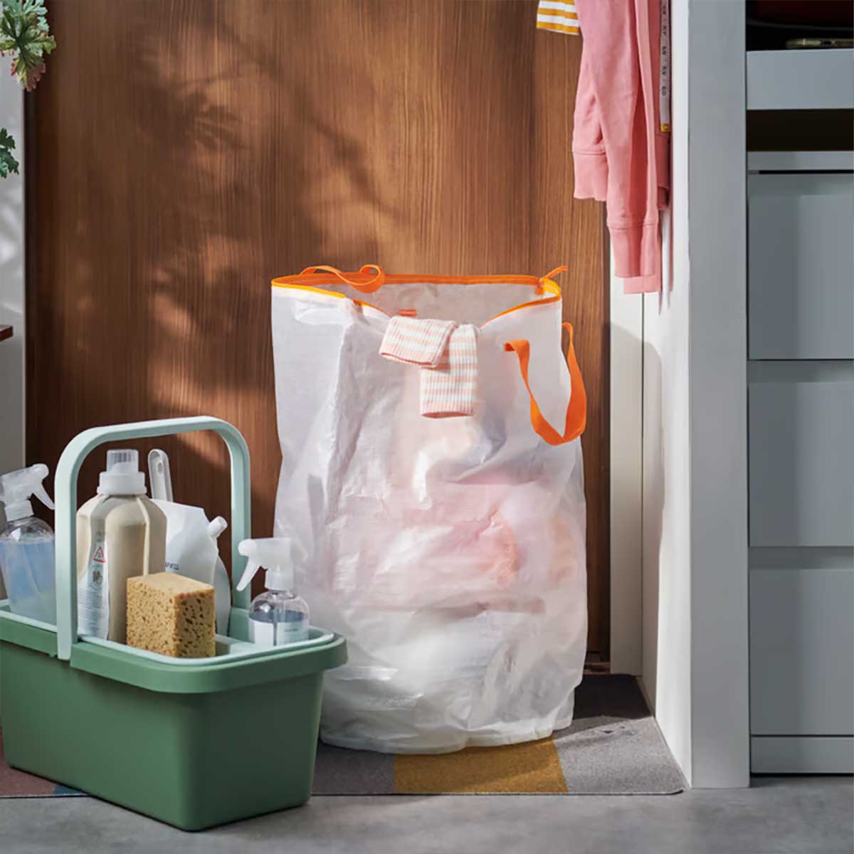 A hallway with a green PEPPRIG cleaning bucket and caddy beside a large white PÄRKLA laundry bag filled with clothes.