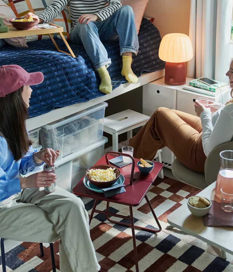 Three students in a dorm room gathered around an IKEA PS 2026 side table, with a KULGLASS speaker lamp in the background.