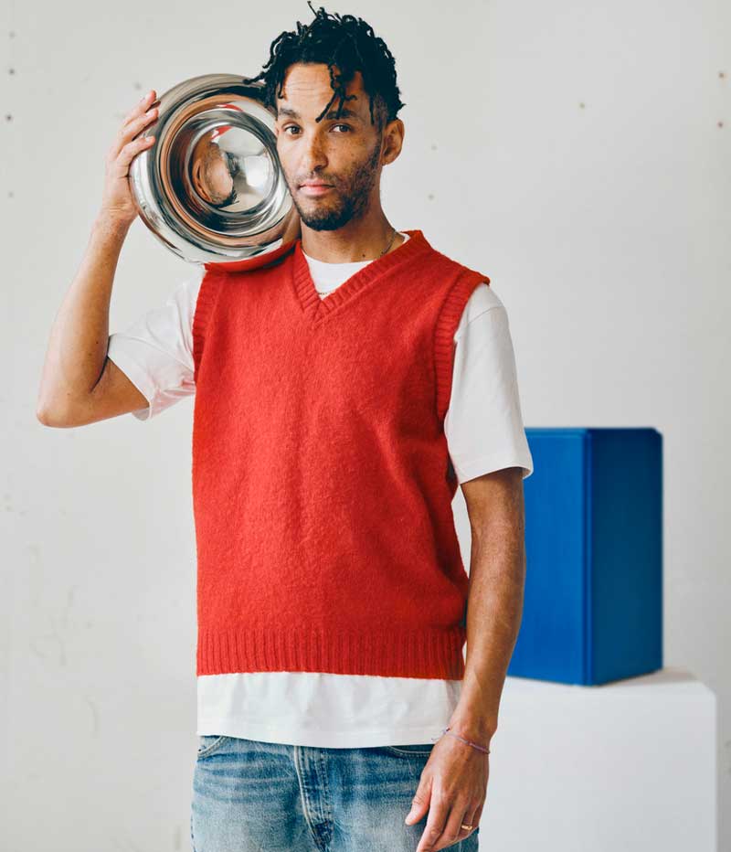 Andu Masebo holding a metallic bowl close to his head, behind him there’s one of his blue sculptures.