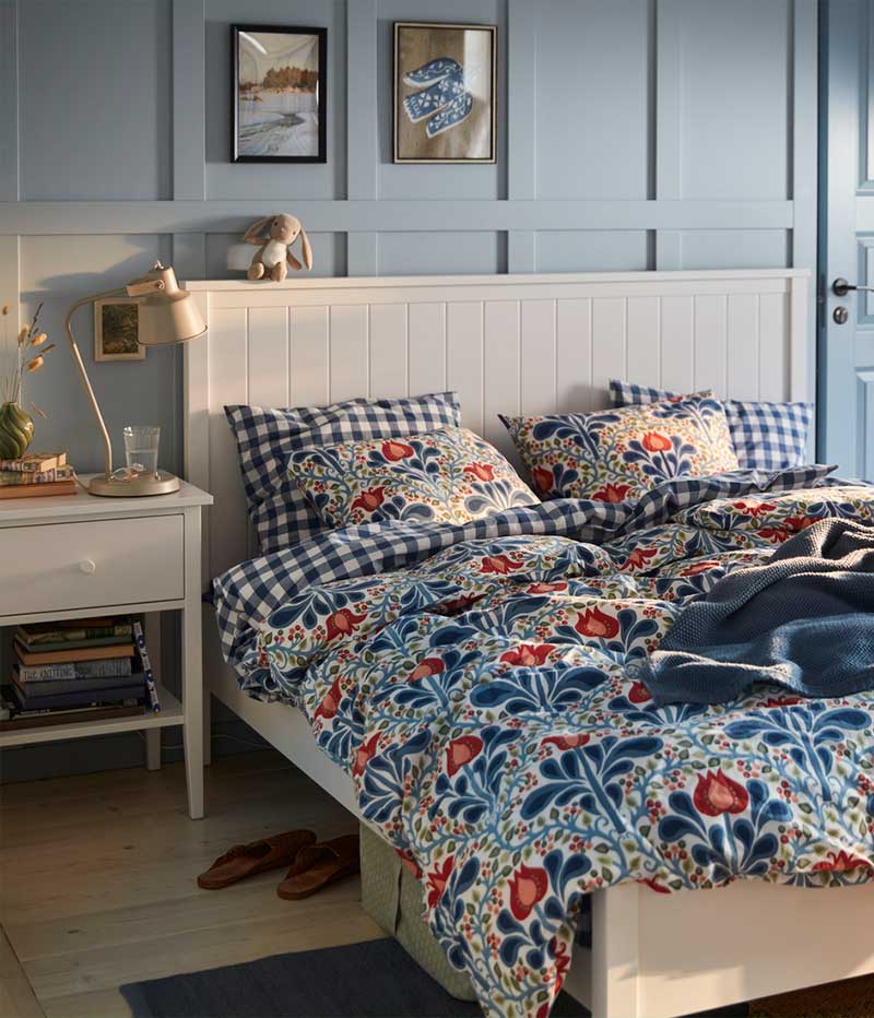 A blue painted bedroom with a floral SÄTERVIDE duvet cover set on a white bed, next to a white bedside table with a lamp.