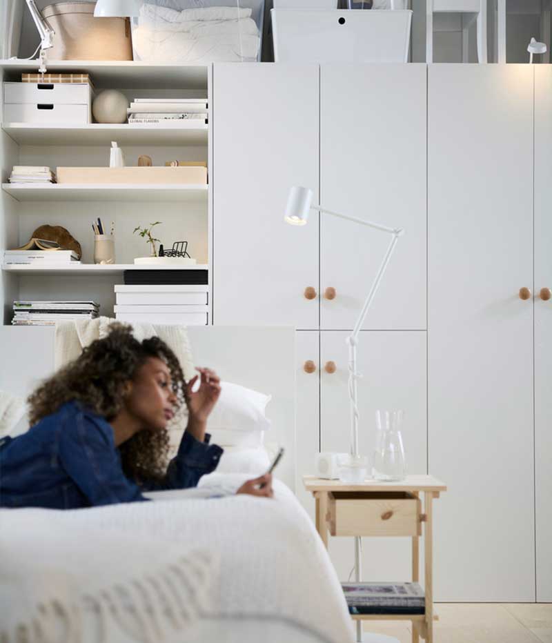 A person relaxes on a bed in a multifunctional living space with white LASTARE cabinets and shelves.