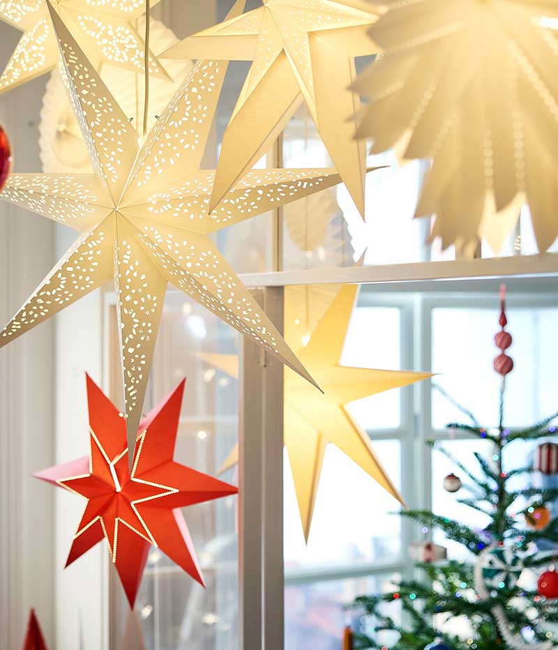 A group of white and red glowing STRÅLA star lampshades hanging from a ceiling with a large red VINTERFINT tree decoration.