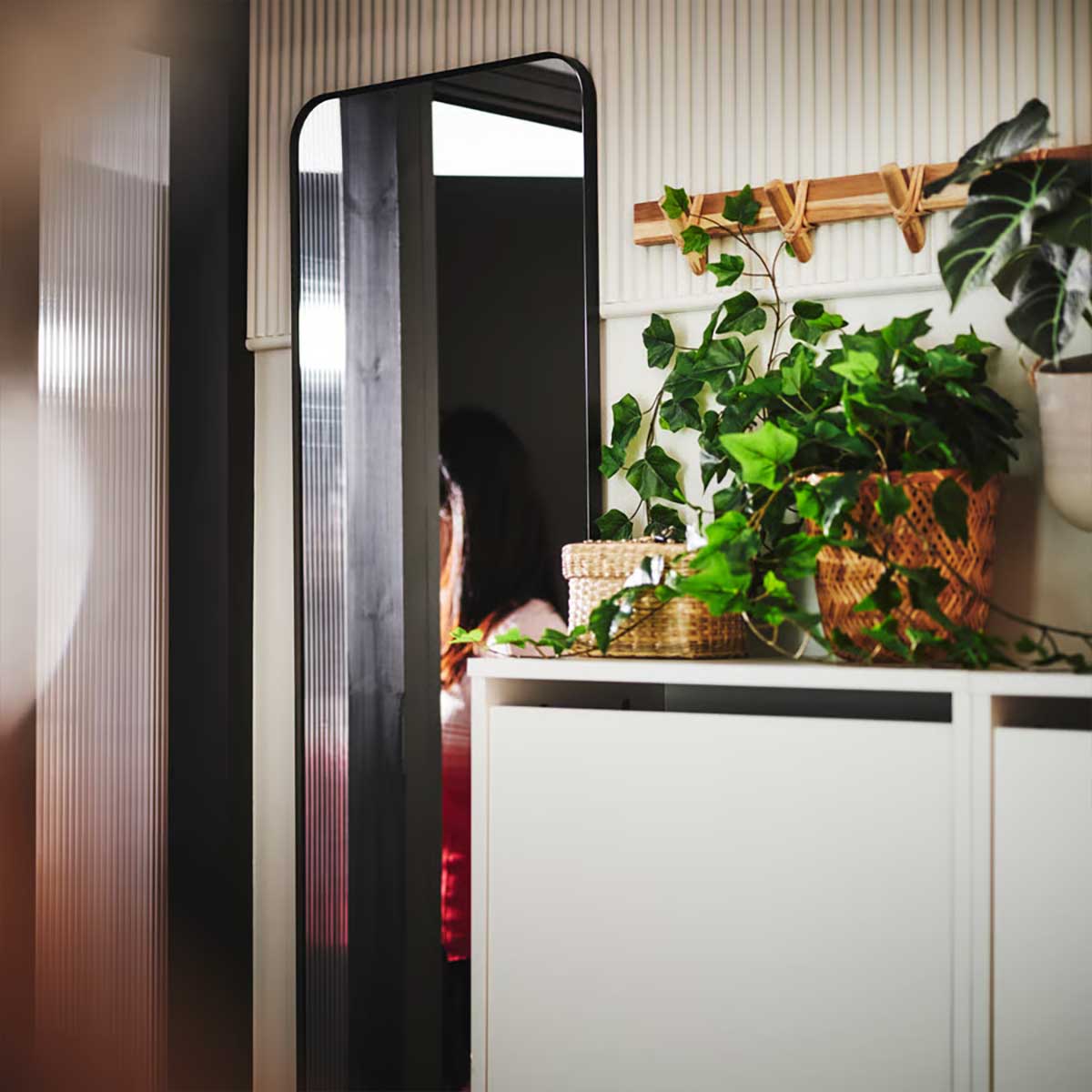 A black LINDBYN mirror by an open front door and two BISSA shoe cabinets with various FEJKA artificial potted plants on top.