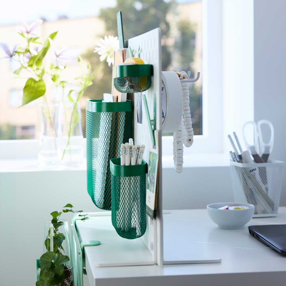 The free-standing SKÅDIS pegboard with  green baskets and clips placed on the edge of  a desk in front of a window.