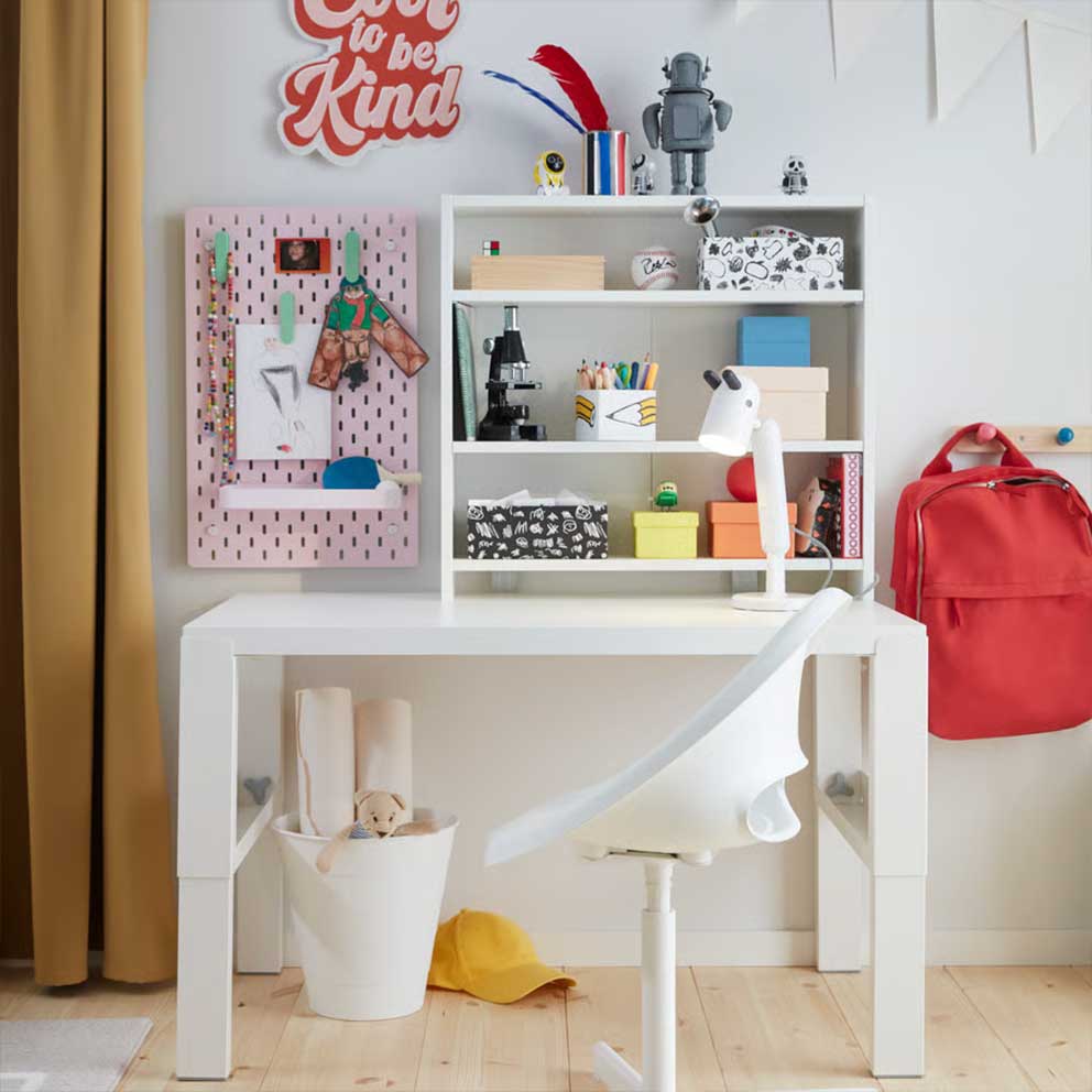 A white PÅHL desk and a white desk chair  placed in a children’s room with a shelf and a  pink SKÅDIS pegboard.
