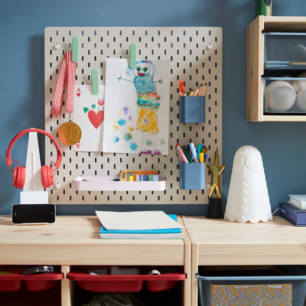 A beige SKÅDIS pegboard with colourful  accessories holding pens and drawings placed  on a blue wall above storage in pine.