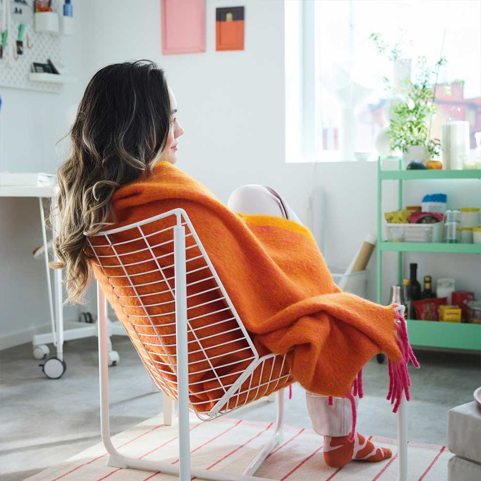 A person sitting in a white net chair wrapped  in the orange KLIPPOXEL throw placed in a  white room with colourful objects.
