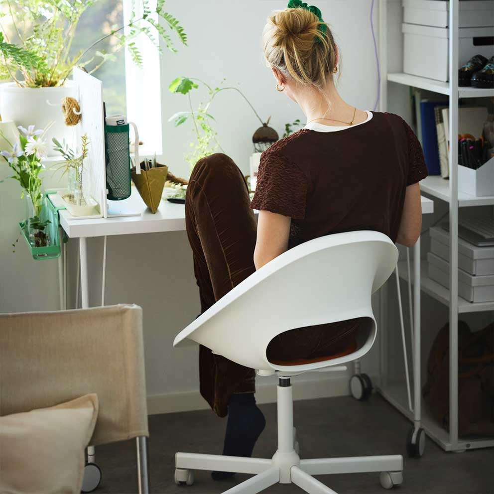 A person dressed in brown sitting on a white  LOBERGET swivel chair by a white desk with  green plants by a window.