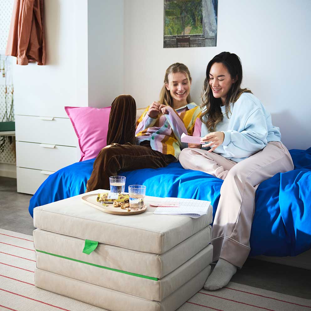 Two students sitting talking in a bed with a  SLÄKT pouffe in front of them holding a tray  with snacks and water.