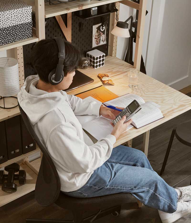 A teenager studying by an IVAR folding table  in pine attached to an IVAR shelving unit  holding black and white objects.