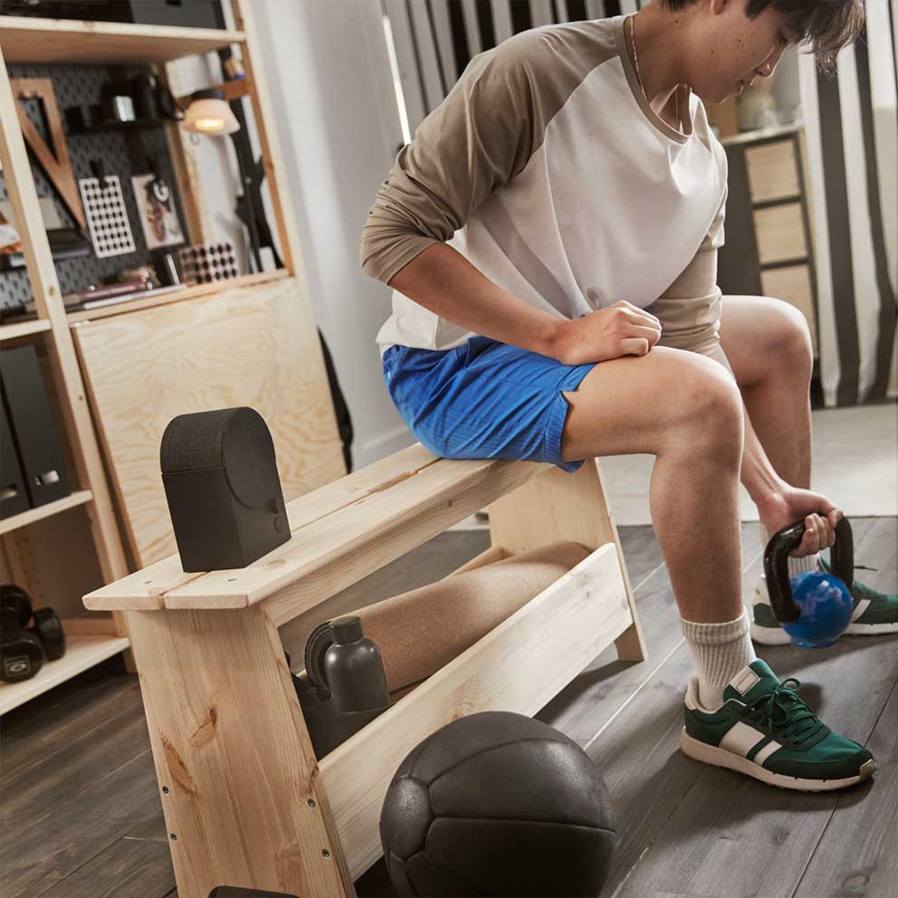 A black NATTBAD Bluetooth speaker with a  rounded shape is standing on a pine bench  where a person is lifting weights.