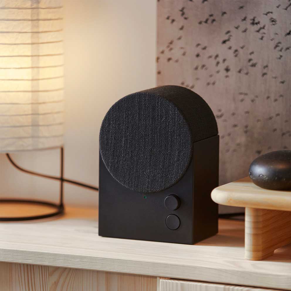 The black NATTBAD Bluetooth speaker with a  rounded shape and two buttons on the front  is standing on top of a pine cabinet.