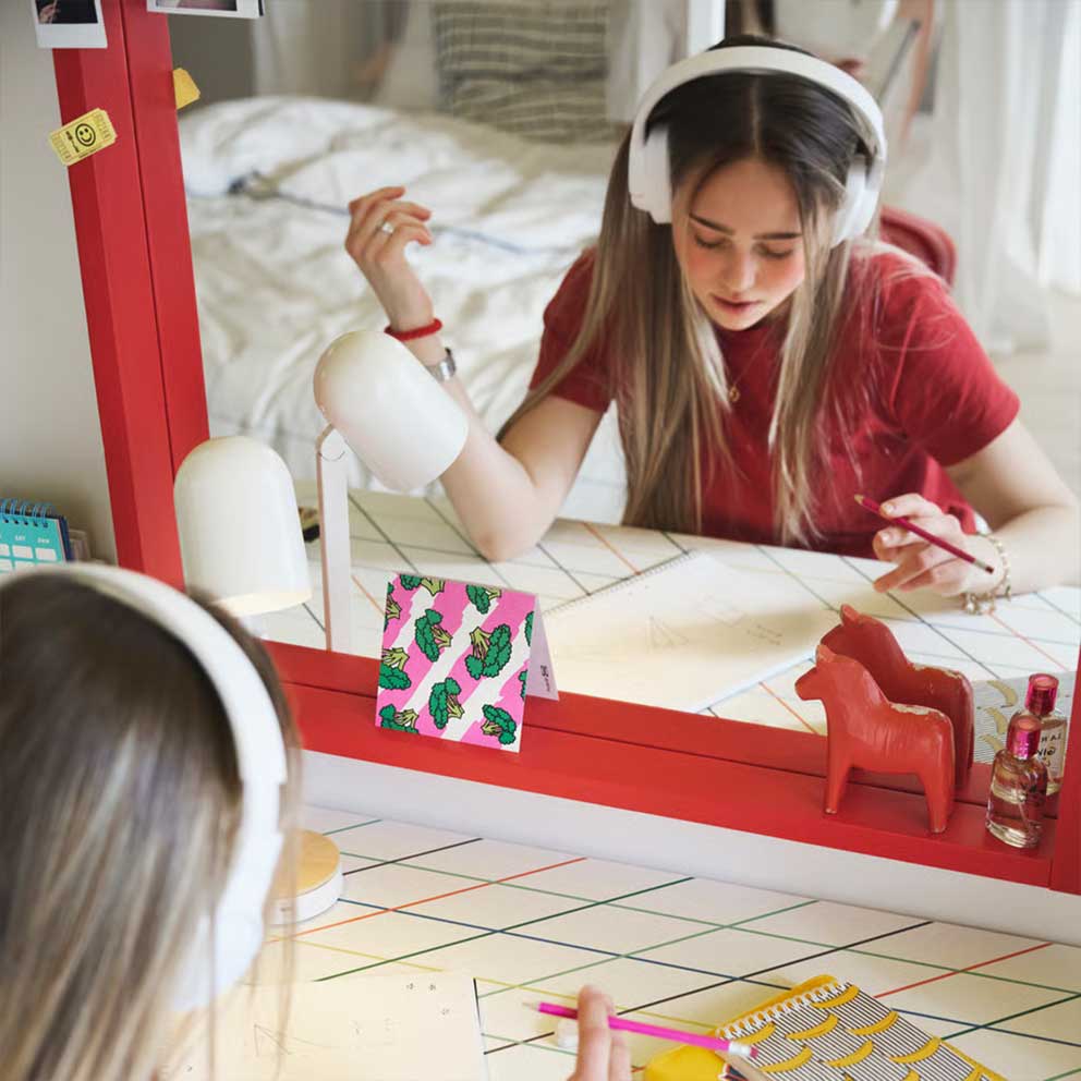 A teenager sitting studying by a LAGKAPTEN/ SPÄND desk with a colourful grid pattern  placed in front of a big red mirror.