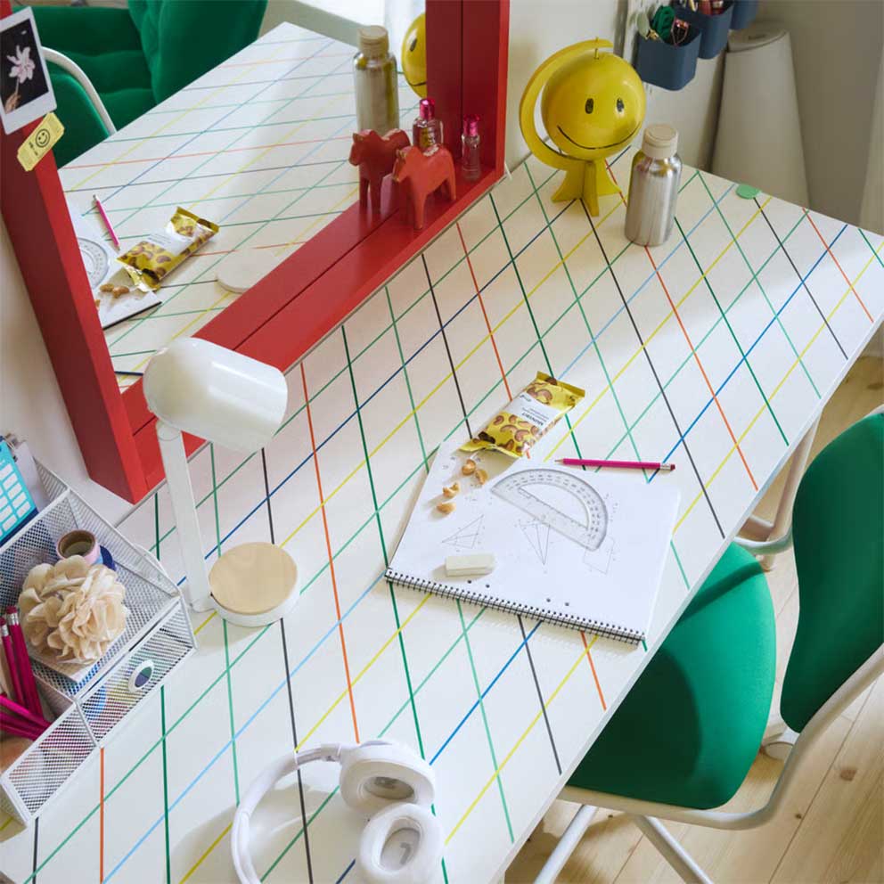 A LAGKAPTEN/SPÄND desk with a colourful  geometric grid pattern paired with a green  desk chair, white lamp and red mirror.