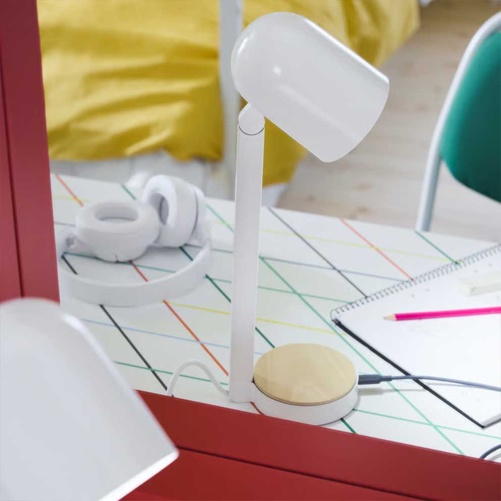 The white ENSMÄRKE desk lamp placed on a  desk with a graphic pattern with a bed in the  background made with yellow bedlinen.