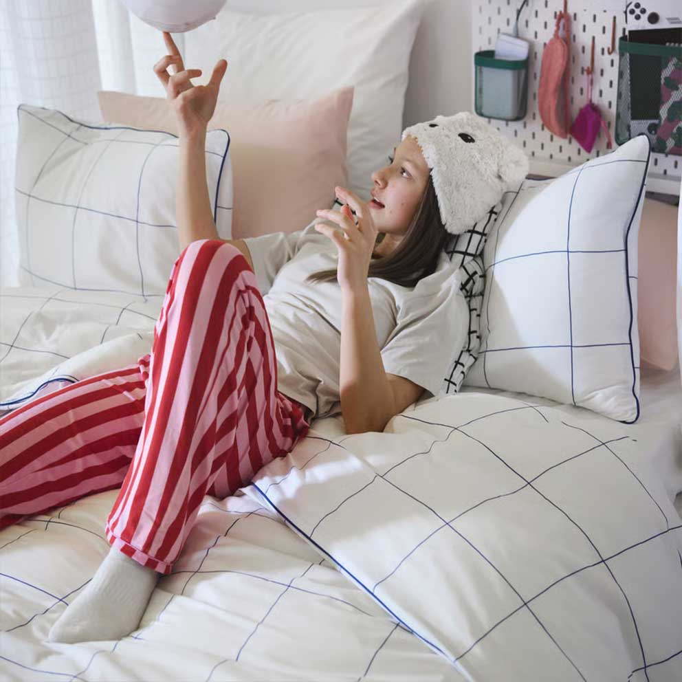 A teenager playing with a round object lying  in a bed made with a BREDVECKLARE duvet  cover in white with a blue grid pattern.